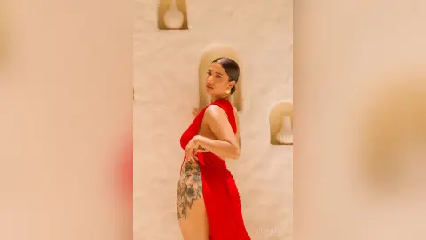 JuliahLewis poses in a red dress with a high slit, showing a large floral tattoo on her thigh against a white wall.