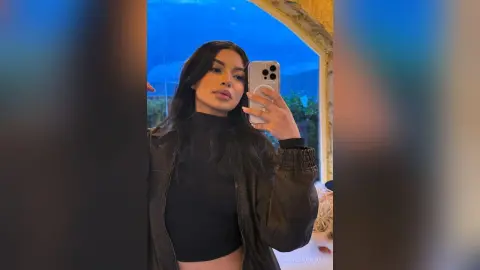 Young woman in a black turtleneck and leather jacket taking a moody mirror selfie against a blue sky background.