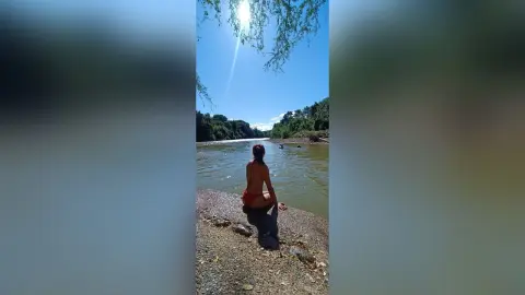 AmelieLautrec sits on a riverbank in red bikini bottoms, showing her bare back and hair while looking out over the water under a bright sun.