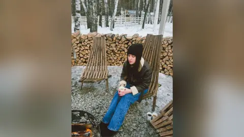 Young woman in a black beanie and jacket relaxing in a wooden chair by a winter bonfire.