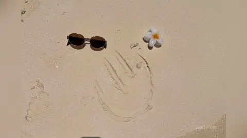 Sunglasses and a white frangipani flower resting on beige sand next to a handprint in a tropical beach setting.