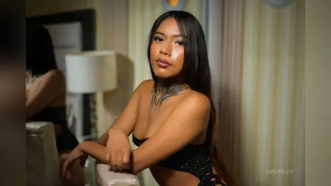 EmaWildy poses with long dark hair, wearing a black cut-out bra or top while leaning against a white chair.