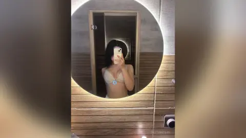 RittaMyers takes a mirror selfie wearing a white bikini top while standing in a wood-paneled shower stall.