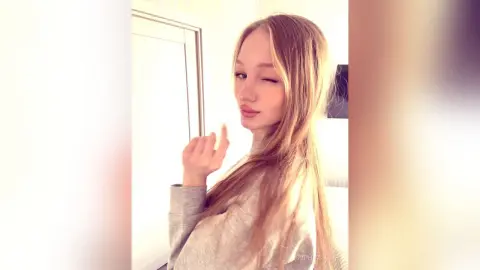 Winking young woman with long blonde hair wearing a grey sweater in a bright indoor setting.