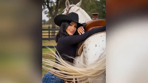 Smiling woman in a black long-sleeve shirt hugging a white horse in a rustic outdoor ranch setting.