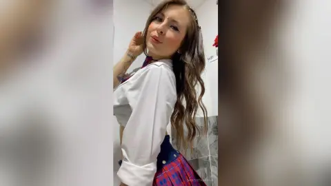 Young woman with long wavy hair wearing a white button-down shirt and plaid skirt in a bright indoor setting.