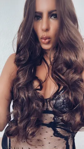 Woman with long wavy brown hair wearing a black lace polka dot bodysuit in a bright indoor setting.
