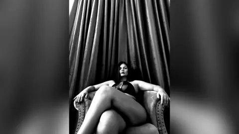 Elegant woman in black lingerie relaxing in a chair against dramatic draped curtains in a moody black and white portrait.