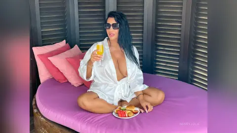 MissyJolie sits cross-legged on a purple bed wearing a white silk robe while holding a glass of orange juice.