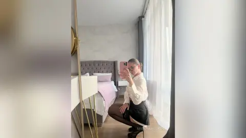 MilanaVolkova poses in a white blouse and black skirt, taking a mirror selfie in her bedroom near a bed and window.