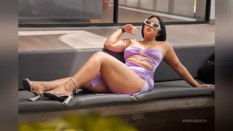 KimRoberth reclines on a grey bench wearing a light purple cut-out bodysuit and white sunglasses.