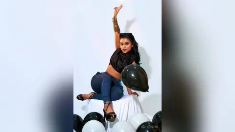 Cheerful young woman in a black crop top and blue jeans posing with black balloons against a white background.