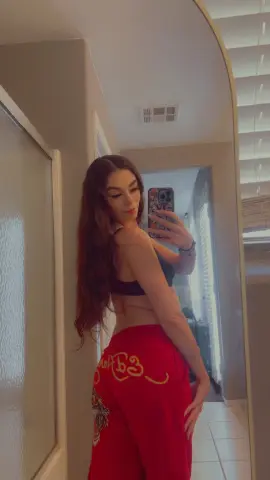 Young woman in black sports bra and red graphic sweatpants taking a mirror selfie in a bright bedroom.