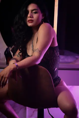 Moody portrait of a woman with long dark hair wearing a black lace bodysuit in a dimly lit room.