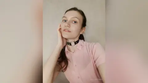 Young woman in a pink polo shirt and black choker posing with a soft expression against a neutral wall.