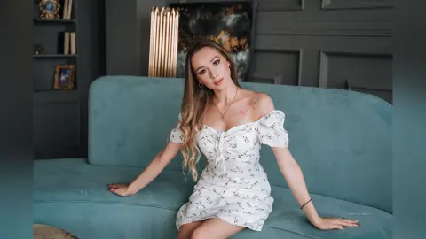 Elegant woman in a white floral sundress sitting on a teal velvet sofa in a moody, modern living room.