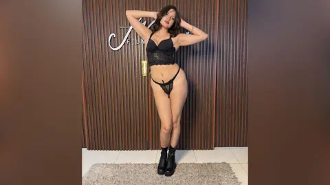 Young woman in black lace lingerie and boots posing confidently against a wood-paneled wall.