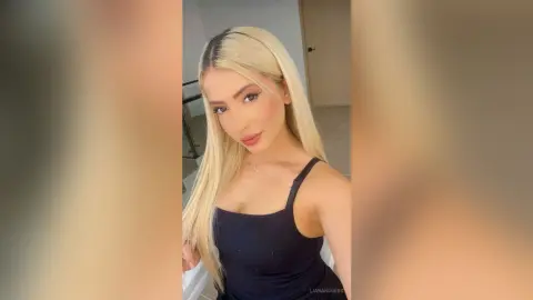 Blonde woman in a black tank top posing for a selfie in a bright, modern indoor setting.