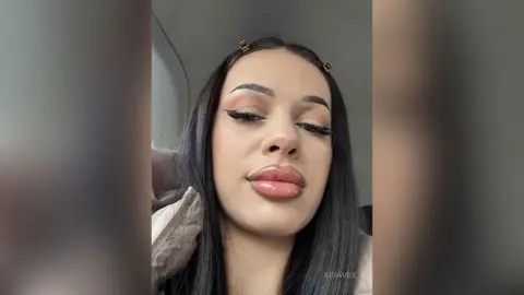AriaVex looks directly at the camera with long dark hair and heavy eyeliner, wearing a light tan jacket.