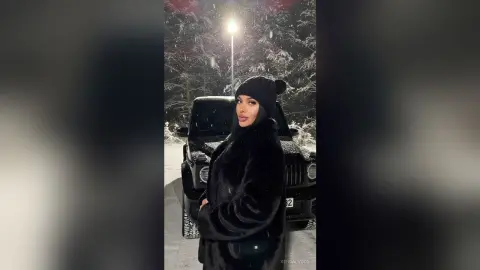 Stylish woman in a black faux fur coat and beanie posing by a Jeep in a snowy winter forest at night.