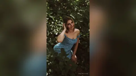 Smiling young woman in a blue floral sundress relaxing in a lush green garden setting.