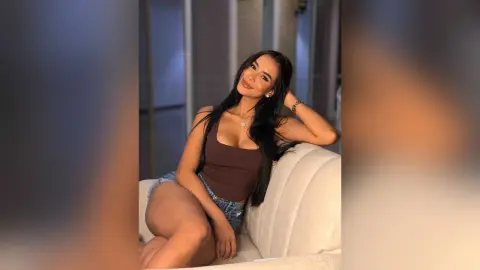 Alison sits on a white sofa wearing a brown tank top and denim shorts, leaning back with one hand in her long dark hair.