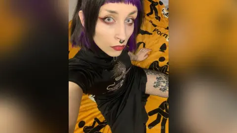 Woman with purple hair and septum piercing wearing a black graphic t-shirt in a moody indoor setting.
