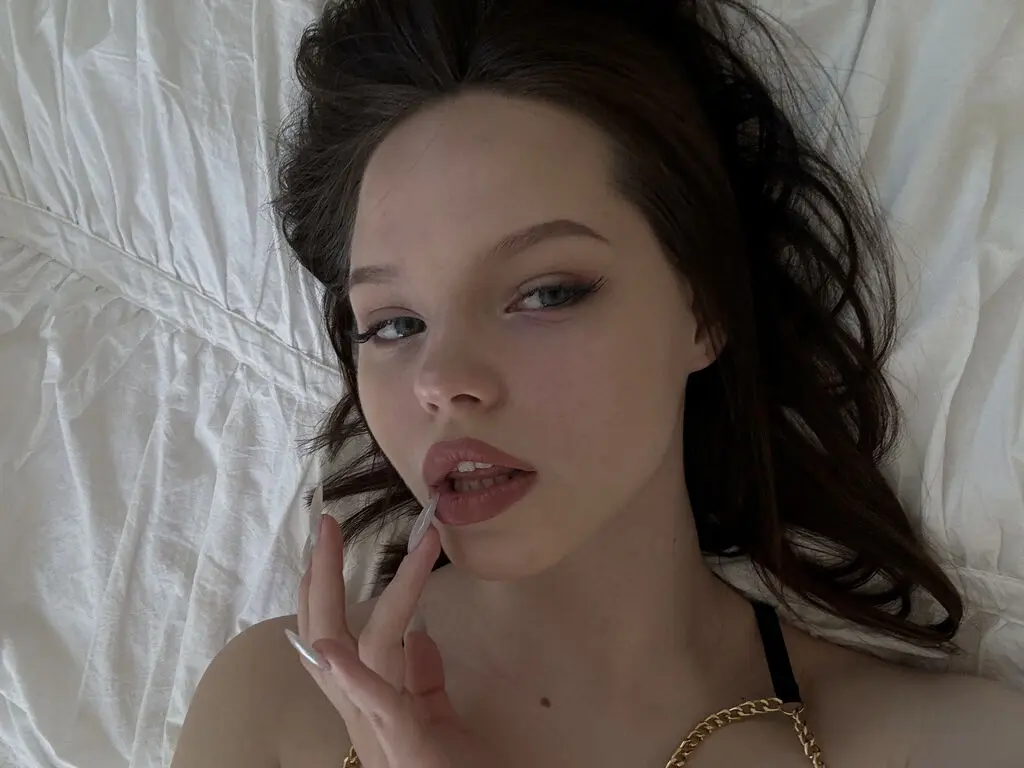 SelenaNoelle lies on white bedding with her dark hair spread out, looking at the camera with long nails near her lips.