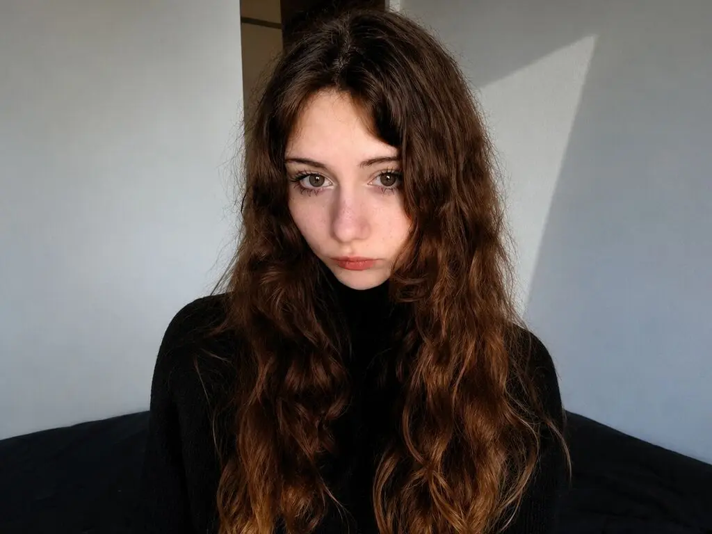 Young woman with long wavy brown hair wearing a black turtleneck in a minimalist indoor setting.
