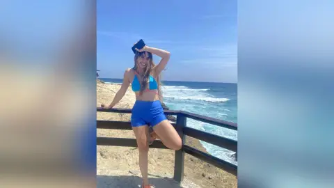 Smiling woman in blue bikini top and shorts posing on a wooden pier overlooking the sunny ocean waves.