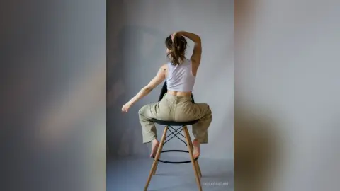 SamaraAdam sits on a stool in a white crop top and tan pants, posing with her hand in her hair against a plain wall.