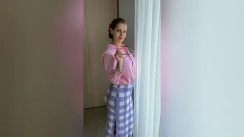 Smiling woman in pink button-down shirt and purple checkered pants standing by a window in a bright room.