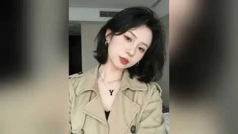 Young woman in a beige trench coat and silver necklace posing with a soft, elegant expression indoors.