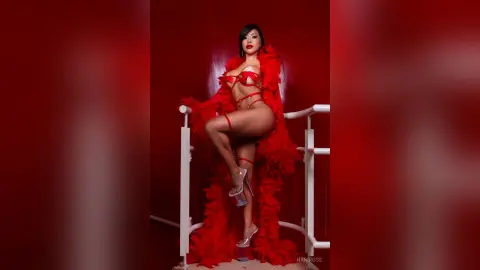 Glamorous woman in red lingerie and feathered robe posing on a white railing against a bold red background.
