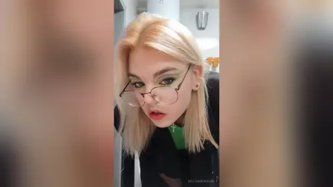 Blonde woman with green eyeshadow and glasses wearing a black outfit in a bright indoor setting.