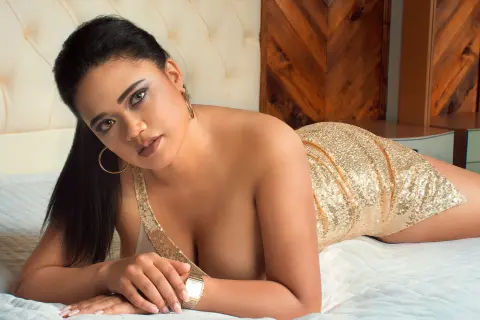 NiaMorelli lies on a bed wearing a gold sequin dress, looking at the camera with her dark hair pulled back.