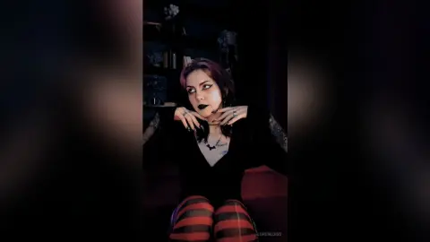 LorenLoiss poses with her hands near her face, wearing black lace, dark lipstick, and red and black striped tights.