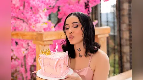 Young woman in a pink top blowing a kiss next to a pink birthday cake amidst blooming pink flowers.