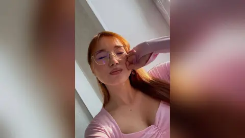 Smiling woman with glasses and long reddish hair wearing a pink top in a bright, airy indoor setting.