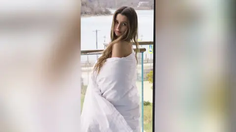 Young woman wrapped in a white blanket posing on a balcony overlooking a serene lake at sunset.