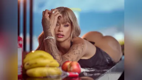 Tattooed blonde woman in black swimwear relaxing outdoors by a bowl of fresh fruit on a sunny day.