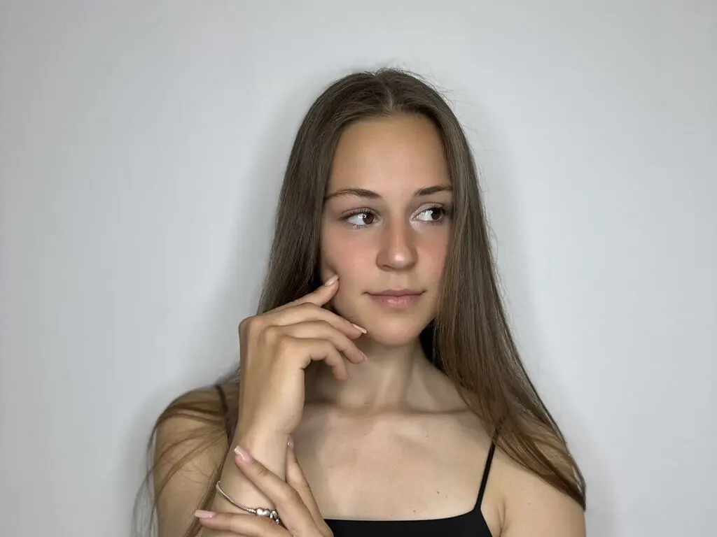 Young woman with long brown hair in a black tank top posing against a white background with a thoughtful expression.