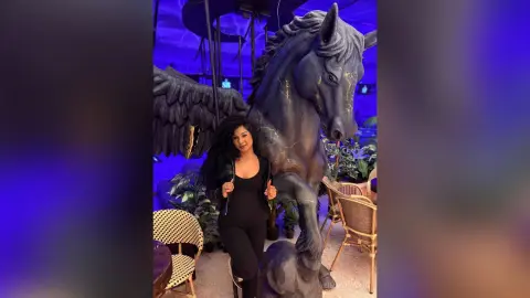 Woman in a black jumpsuit posing next to a large dark winged horse statue in a moody blue-lit lounge.