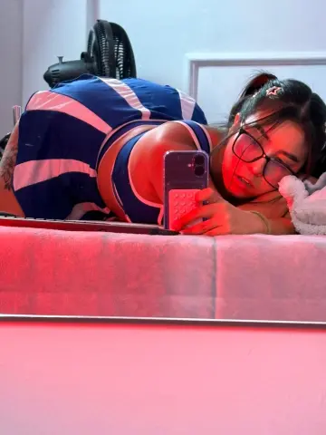 Young woman in a blue striped tank top taking a selfie while lying on a bed in moody pink lighting.