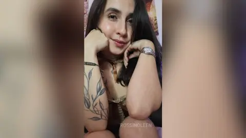 RossiNoleen poses with her chin resting on her hand, showing a leafy vine tattoo on her arm and wearing a brown lace top.