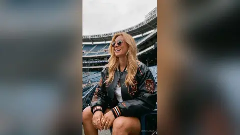 SerenaStar smiles wearing sunglasses and a black and orange letterman jacket while sitting in stadium seating.