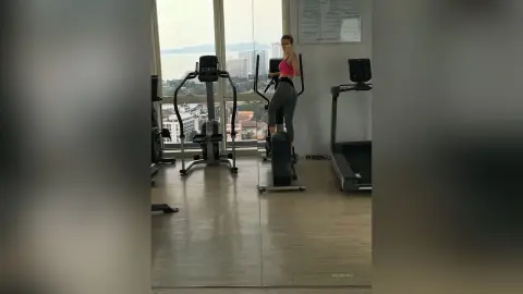 MilenaBia stands in a gym wearing a pink sports bra and grey leggings, looking at the camera near a large window.