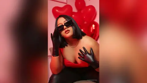 KimRoberth wears black sunglasses and black gloves while posing in a red top against a background of red heart balloons.