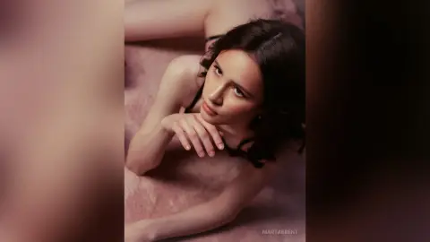Dreamy portrait of a young woman with dark wavy hair resting on soft pink fabric in a moody, intimate setting.