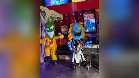 NohomiCambel stands between two large, colorful mascots on a stage with a red background.